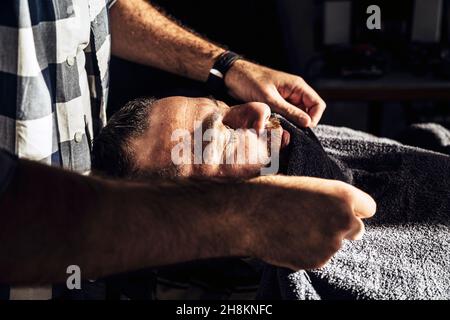 Barbier`s Friseursalon, der das Handtuch dem Kunden ins Gesicht legt. Pre-Shave-Action. Stockfoto